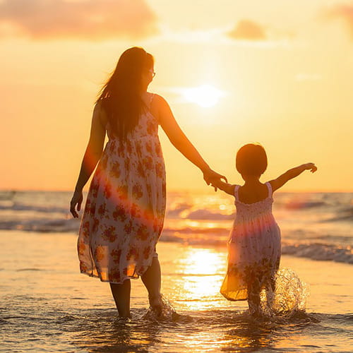 Woman and child holding hands on a beach with the sun setting